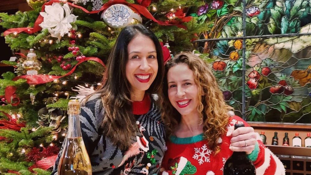 Two women posing and holding wine bottles in front of a Christmas tree